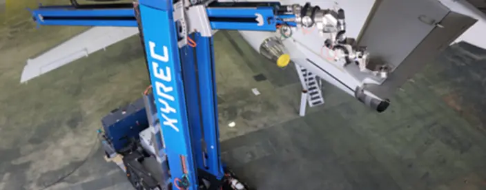 Blue robot comprised of two segments - the first is vertical with XYREC in white lettering; the second is horizontal to the ground with a laser attached. The robot is using a laser to remove an aircraft coating