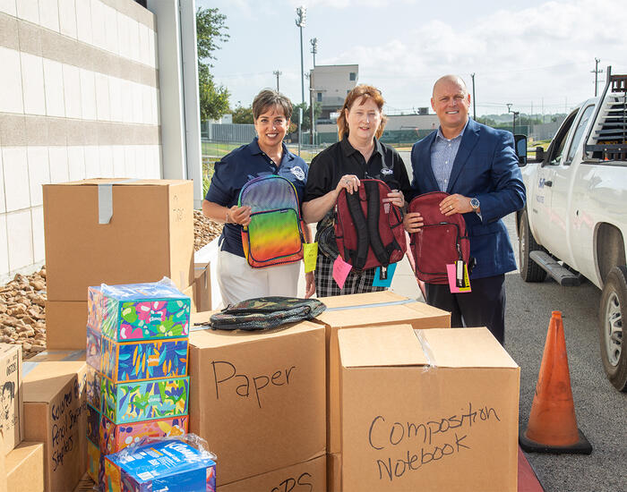 wo women and 1 man holding backpacks standing behind cardboard boxes labeled with types of school supplies 