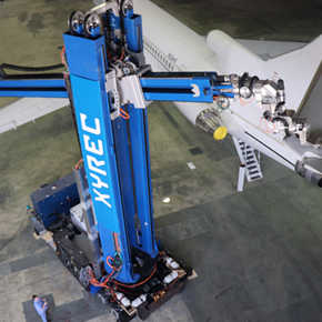 Blue robot comprised of two segments - the first is vertical with XYREC in white lettering; the second is horizontal to the ground with a laser attached. The robot is using a laser to remove an aircraft coating