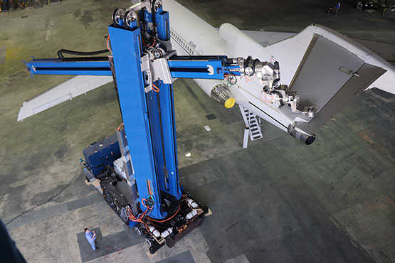 Blue robot comprised of two segments - the first is vertical; the second is horizontal to the ground with a laser attached. The robot is using a laser to remove an aircraft coating