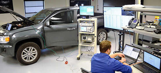 Wires attached to a gray pick up truck with the hood popped up. Wires connect to computer lap station. Engineers identify vulnerabilities on vehicles and in electronics modules. 